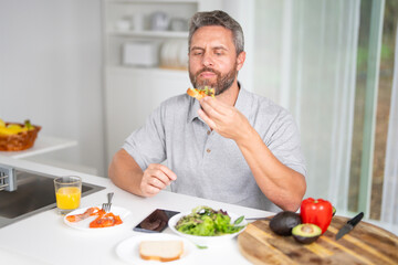 Fototapeta premium Man eat breakfast in the kitchen on a morning. Guy eat vegetables for a healthy meal in his kitchen in the morning. Man eat healthy food in cozy kitchen. Morning breakfast in the modern kitchen.