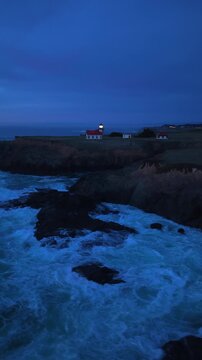 Vertical Screen: Night aerial view of coastal lighthouse buildings with glowing light on rocky cliffs as powerful ocean waves crash against the shoreline during blue hour twilight