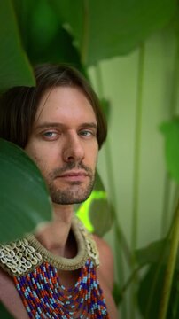 Young Man with Beaded Necklace Looking at Camera Among Tropical Leaves