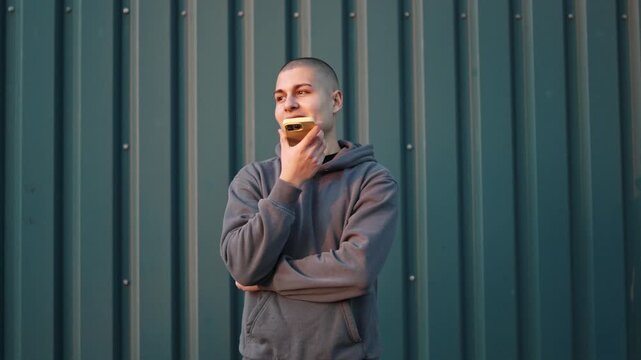 Young man with buzz cut recording a voice message on his smartphone against a corrugated metal wall outdoors