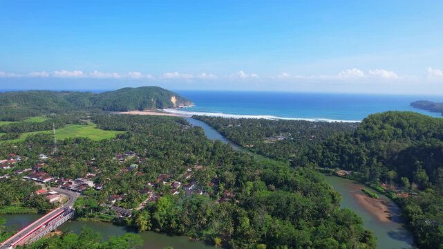 Aerial drone footage of Taman beach bay, in Ngadirojo, Pacitan regency Java island, Indonesia, with a large forest until the beach, a wide river, blue sea water, some local houses, and a bridge