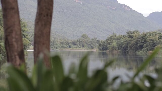 A scenic view of a calm river and lush green mountains seen through trees in Kanchanaburi, Thailand. A tranquil and refreshing tropical landscape.