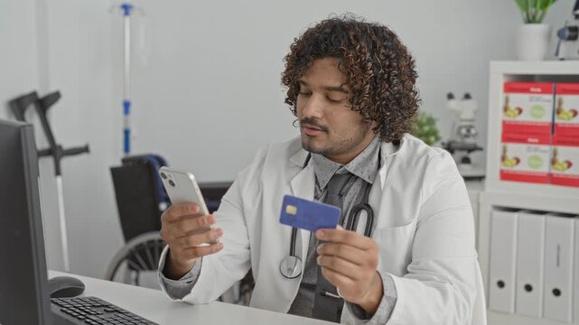 Man doctor with stethoscope smiling while holding smartphone and creditcard at a clinic desk in a clinic building; trust convenience.