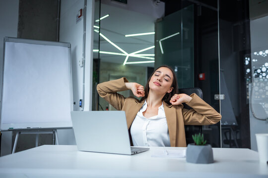 young smiling woman sitting in the office in front of a laptop and happily stretching after work. Brunette woman with closed eyes have a break