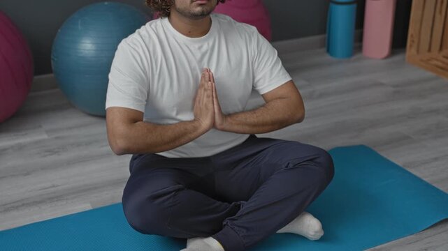 Man seated crosslegged with hands pressed in prayer pose on a yoga mat in studio; mindfulness breathing calm.
