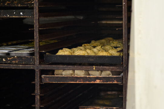 Trays of traditional Maltese pastizzi and wraps inside a professional metal baking oven rack. The pastries are arranged on dark baking sheets.
