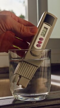 First Person View Of Hand Measuring Purity Of Drinking Water With Digital Tester Showing Twenty Three Ppm In Clear Glass Near Kitchen Sink And Window With Tropical Green Landscape. Water Quality Contr