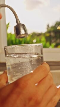 First Person View Of Hand Filling Glass With Tap Water Under Kitchen Faucet Before Measuring Ppm With Digital Tester Near Window With Tropical Green Landscape. Drinking Water Quality Check