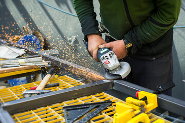 A worker uses a grinder to cut metal in a workshop. Sparks are flying as the tool makes contact with the material. Tools are scattered on the table nearby