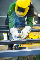 A worker wearing a yellow helmet and gloves carries out welding work on a metal frame in a construction area. Smoke rises as sparks fly, showing the intensity of the task