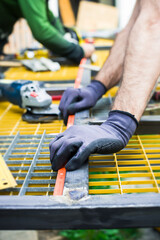 Two workers focus on measuring and cutting metal parts in a workshop. They use tools while ensuring accuracy on a yellow work surface. The setting is bright and active
