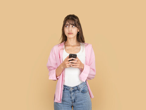 Confused caucasian woman holding smartphone and looking aside with doubtful expression, taking selfie moment, wearing pink shirt and denim, isolated on beige studio background with copy space.