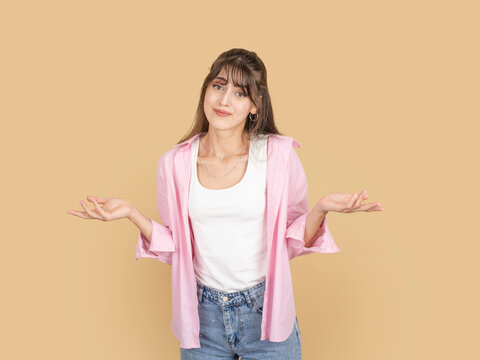 Confused caucasian woman shrugging shoulders with unsure expression, reacting to unclear situation, taking selfie mood, wearing pink shirt and denim, isolated on beige background with copy space.