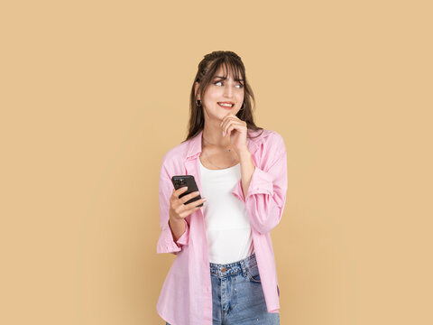 Smiling caucasian woman holding smartphone and touching chin while thinking, looking aside with positive expression, wearing pink shirt and denim, isolated on beige studio background with copy space.
