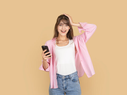 Surprised caucasian woman holding smartphone and touching head with amazed expression, reacting to unexpected message, wearing pink shirt and denim, isolated on beige background with copy space.