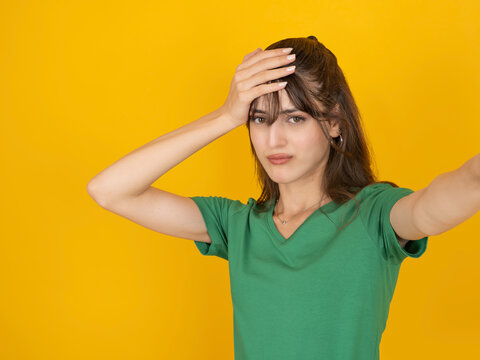 Worried caucasian woman taking selfie and holding her head with stressed expression, reacting to mistake or problem, wearing green t shirt, isolated on bright yellow studio background with copy space.