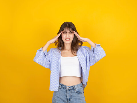 Stressed caucasian woman holding temples with painful expression, experiencing headache or mental overload, wearing striped shirt and denim, isolated yellow studio background with copy space.