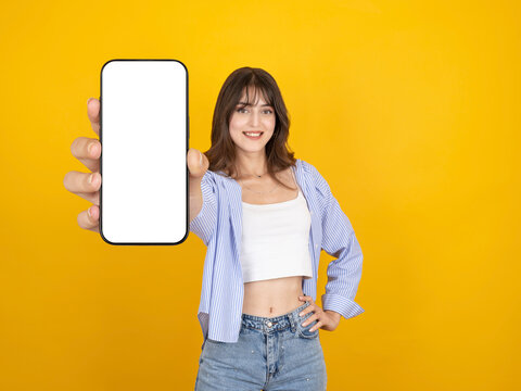 Smiling caucasian woman holding smartphone toward camera with blank screen for mock up, presenting app or content, wearing striped shirt and denim, isolated yellow studio background with copy space.