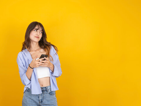 Caucasian woman holding smartphone and looking aside with suspicious expression, reacting with doubt or hesitation, wearing striped shirt and denim, isolated yellow studio background with copy space.