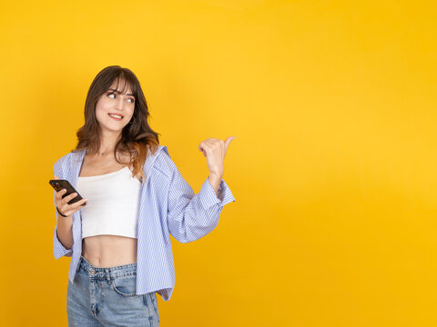 Smiling caucasian woman holding smartphone and pointing behind with thumb, suggesting or recommending something, wearing striped shirt and denim, isolated on yellow studio background with copyspace.