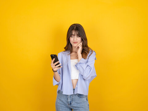 Thoughtful caucasian woman looking at smartphone with slight doubt, touching her chin while thinking, wearing striped shirt and denim, isolated on bright yellow studio background with copy space.