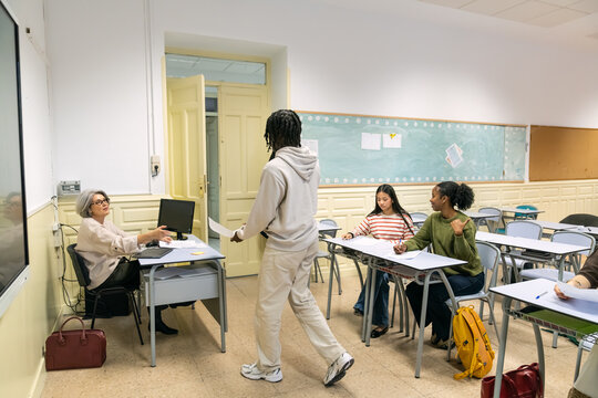 Diverse students in a high school or college classroom, one student submitting a test to the teacher after finishing