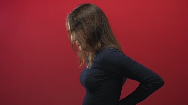 Woman holding lower back in red studio with head bowed and hair covering face, facing sideways; pain solitude.
