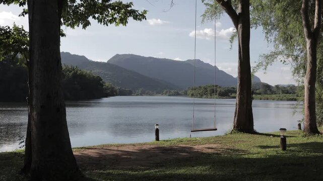 A beautiful morning timelapse of a calm river with a wooden swing and lush mountain reflections in Kanchanaburi, Thailand. Serene and tranquil tropical landscape.
