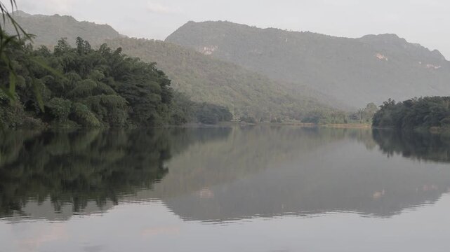 A stunning view of lush green mountains perfectly reflecting on the mirror-like surface of a calm river in Kanchanaburi, Thailand. Serene morning scenery.