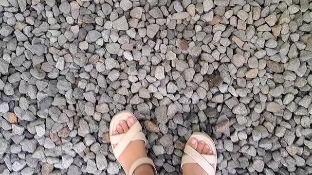 A horizontal first-person perspective looking down at feet in white sandals on a pebble path.