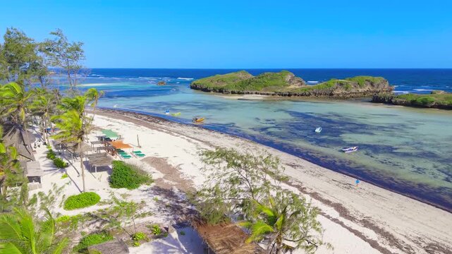 Aerial view of thatched roof huts and palm trees along the white sand coastline with rocky islands and clear water at Watamu Beach, Kenya.