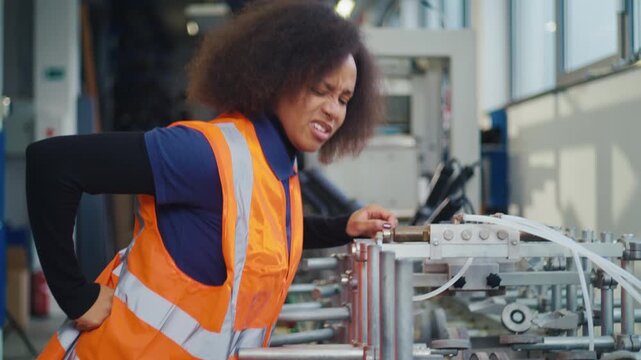 African American female industrial worker having physical workload and back pain while checking machinery, workplace injury in manufacturing