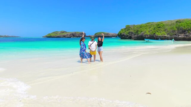 Aerial shot of two young women and a young man standing in shallow turquoise water on a white sand beach in Watamu, Kenya.