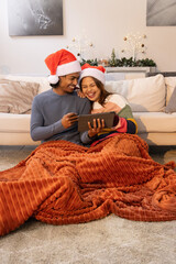 Diverse couple wearing Santa hats, leaning close on rug, sharing tablet and rust-orange blanket © wavebreak3