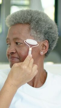 Vertical video: Placing pink roller on senior cheek by window, aide rolling for care, white shirt