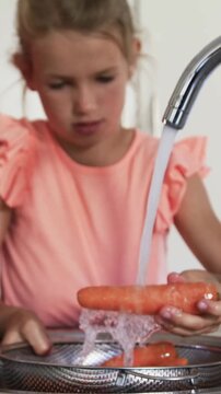 Turning faucet, child wearing coral top rinsing carrots at kitchen sink with colander for cooking