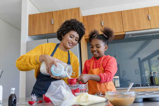 Pouring flour from jar into red measuring cup by diverse mother and daughter in home kitchen