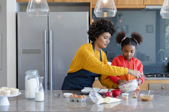 African American mom and daughter mixing batter at kitchen island, with mixing bowl, measuring cups