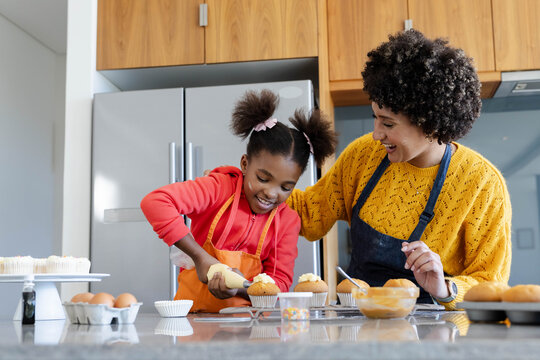 African American mother and daughter frosting cupcakes in kitchen, with piping bag, sprinkles