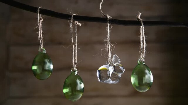 Glass Easter decorations transparent bunny and three ribbed green eggs are suspended on rustic-style twine from dark branch against a warm wooden background, creating atmosphere of cozy spring decor