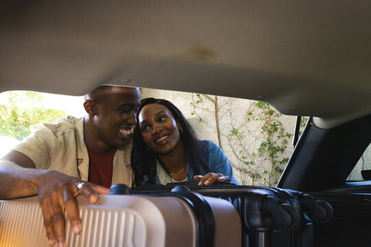 African American couple leaning into car trunk on sunny driveway, loading two rolling suitcases