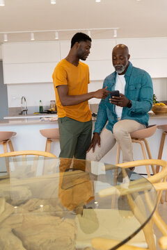 African American father and son sharing smartphone tips in modern kitchen, with fruit bowl