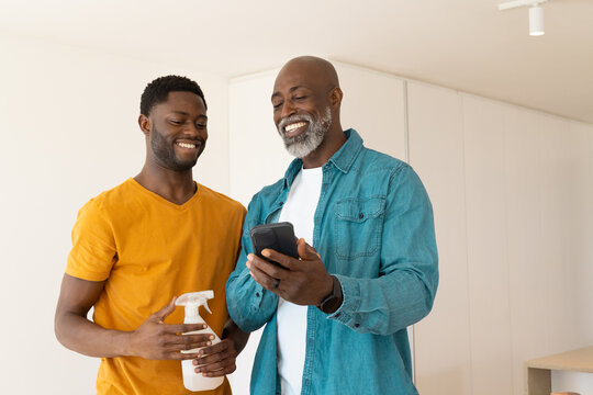 African American father and son using phone and spray bottle in kitchen, sharing cleaning task