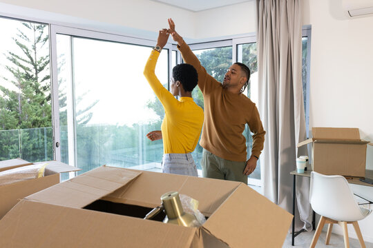 Diverse couple dancing in living area near sliding glass doors, surrounded by unpacked boxes