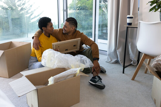 Sitting diverse couple unpacking cardboard boxes on floor at home, with unframed picture frame