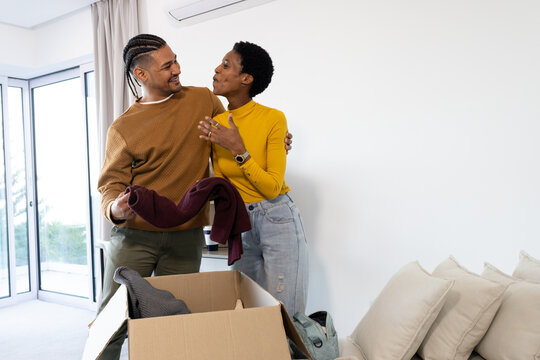 Unpacking diverse couple hugging in living room, holding folded sweater from cardboard box