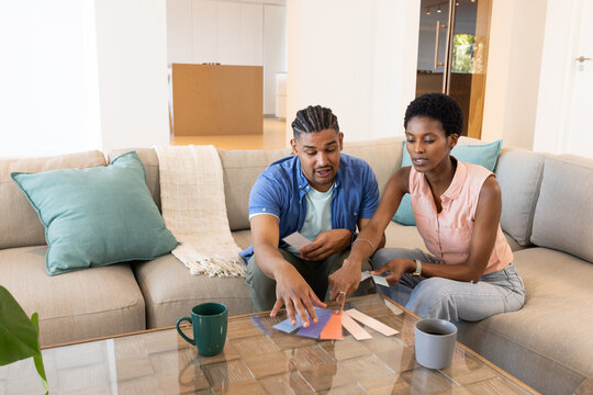 Diverse couple leaning forward over coffee table in living room, with paint sample cards