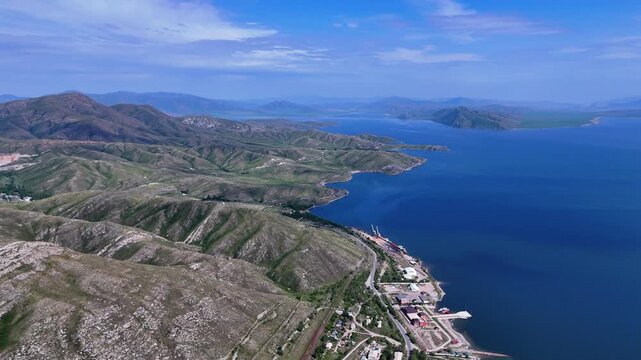 Stunning drone footage of the immense Bukhtarma Reservoir in East Kazakhstan. Expansive turquoise waters stretch across the frame, framed by rolling hilly shores and a quaint village nestled on the ba