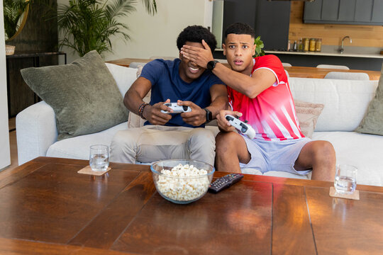 Playing Diverse friends holding game controllers on sofa in living room, with popcorn and water