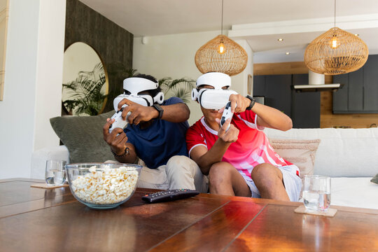 Diverse male friends playing virtual reality games on couch at home, with white VR headsets